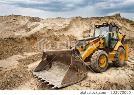 A yellow backhoe loader works on a construction site, moving dirt and debris while clouds gather ominously overhead, signaling possible rain 132670000