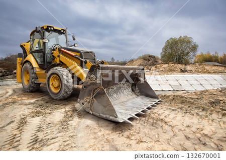 A yellow backhoe loader is smoothing out the ground in a rural area. The cloudy sky hints at possible rain, adding a dramatic feel to the construction site 132670001
