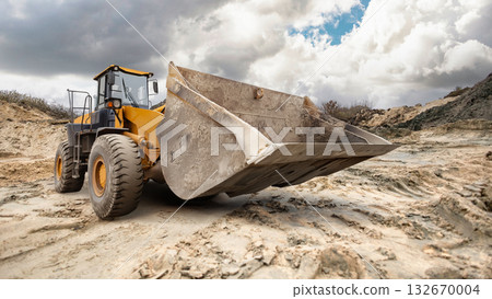 A large excavator loader digs into the sandy terrain at a construction site, surrounded by clouds softly drifting across the blue sky. 132670004