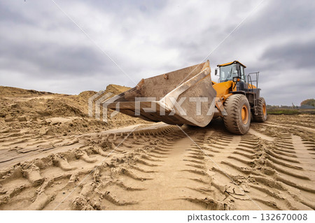A large yellow bulldozer works diligently on a cloudy day, shaping the soil at a construction site surrounded by piles of dirt and tire marks on the ground 132670008
