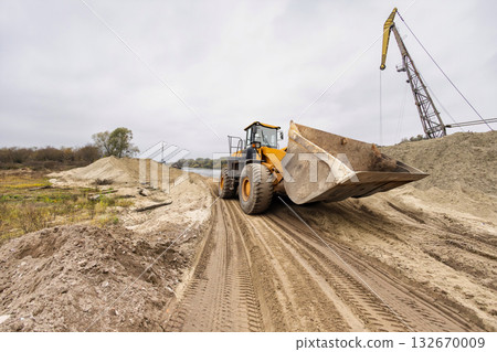 A giant loader works diligently, shifting soil along a riverbank, surrounded by mounds of dirt under a cloudy sky, signaling ongoing construction activity A giant loader works diligently, shifting soil along a riverbank, surrounded by mounds of dirt under a cloudy sky, signaling ongoing construction activity 132670009