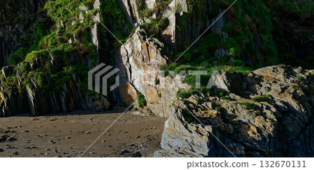 This is a view of the shoreline on a sunny day in West Cork. The rocks form dramatic cliffs with green plants growing on them, and the sand is visible at the base. This is a view of the shoreline on a sunny day in West Cork. The rocks form dramatic cliffs with green plants growing on them, and the sand is visible at the base. 132670131
