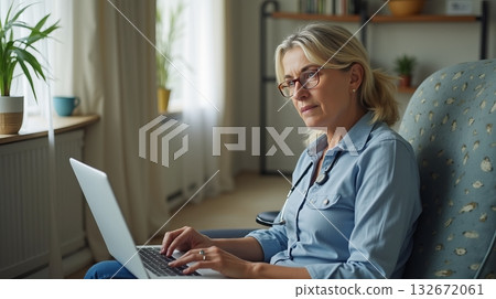 Middle-aged woman wearing glasses and a stethoscope, working on a laptop at home. She sits in a cozy chair with a thoughtful expression 132672061