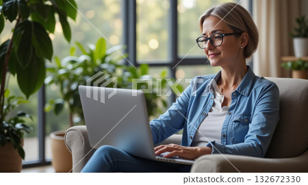 Smiling woman in glasses using a laptop while sitting in a cozy chair, surrounded by green plants near a large window. Concept of remote work 132672330