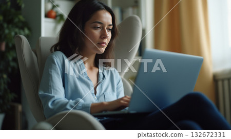 Young woman in blue shirt sitting in cozy chair using laptop in well-lit home interior. She looks concentrated, working remotely or studying Young woman in blue shirt sitting in cozy chair using laptop in well-lit home interior. She looks concentrated, working remotely or studying 132672331