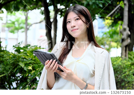 一位年輕的女商人正在公園的綠蔭下使用平板電腦。 132673150