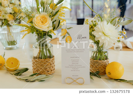 Table setting featuring a menu card with lemons, surrounded by elegant floral arrangements in glass vases. Fresh lemons and greenery add a touch of color and freshness to the table 132675578