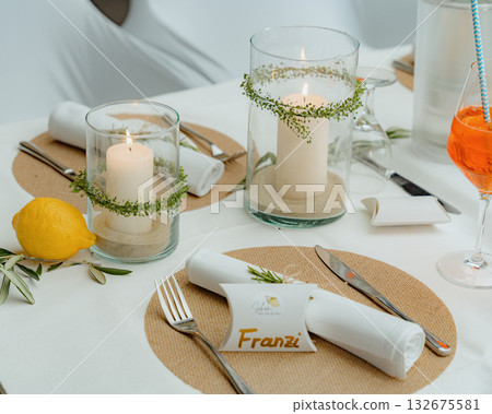 Elegant table setting featuring lit candles in glass holders, a fresh lemon accent, and neatly folded napkins. Ideal for weddings, intimate gatherings, or special events Elegant table setting featuring lit candles in glass holders, a fresh lemon accent, and neatly folded napkins. Ideal for weddings, intimate gatherings, or special events 132675581