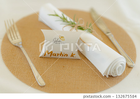 Elegant table setting features a personalized name card, silver cutlery, and a rolled white napkin adorned with rosemary on a woven placemat, creating an inviting dining atmosphere 132675584