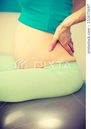 Pregnant woman sitting on fit ball holds hand on her back Pregnant woman sitting on fit ball holds hand on her back 132675697