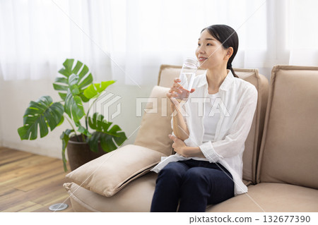 A middle-aged woman sitting on a sofa and smiling while holding a bottle of water 132677390