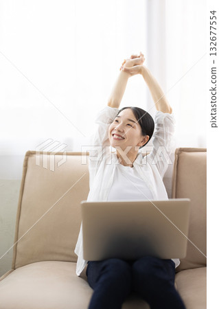一位中年婦女躺在沙發上伸懶腰，膝上放著筆記型電腦，臉上帶著微笑。 132677554