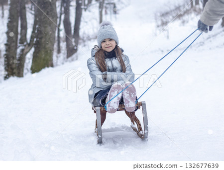 Young girl on a wooden sled being pulled through a snowy forest by a parent in winter joy 132677639