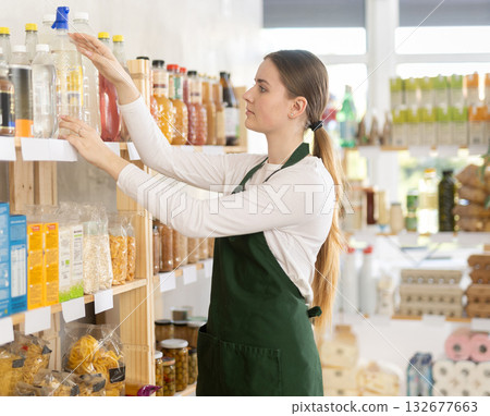 Young woman seller in apron stands in trading hall of store Young woman seller in apron stands in trading hall of store 132677663
