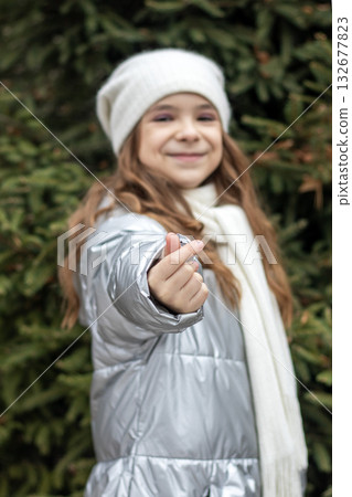 Young girl in silver puffer jacket and white beanie smiles and points at camera outdoors 132677823