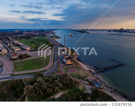 Aerial view of coastal zone at sunset curved shoreline, helipad peninsula, ship in golden water, wind turbines on horizon, and residential grid blend maritime activity with renewable energy. Aerial view of coastal zone at sunset curved shoreline, helipad peninsula, ship in golden water, wind turbines on horizon, and residential grid blend maritime activity with renewable energy. 132679558