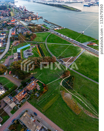 Aerial view of coastal urban recreational zone road with vehicles, red bike path, circular lookout, geometric green fields, tennis courts, and dense residential blocks framed by forest and shoreline. 132679560