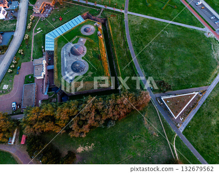 Aerial view of coastal urban recreational zone road with vehicles, red bike path, circular lookout, geometric green fields, tennis courts, and dense residential blocks framed by forest and shoreline. Aerial view of coastal urban recreational zone road with vehicles, red bike path, circular lookout, geometric green fields, tennis courts, and dense residential blocks framed by forest and shoreline. 132679562