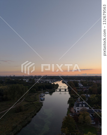 Aerial sunrise view of tranquil river town soft orange pink sky reflects on calm water, bridge connects lush banks, boats docked near homes, blending nature with peaceful habitation. Aerial sunrise view of tranquil river town soft orange pink sky reflects on calm water, bridge connects lush banks, boats docked near homes, blending nature with peaceful habitation. 132679563