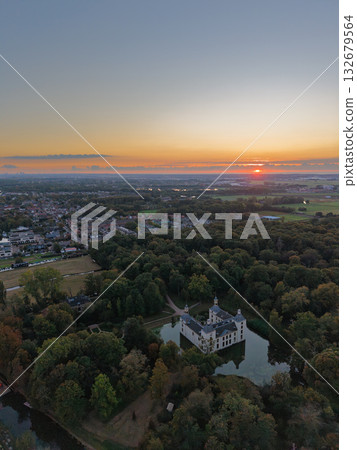 Aerial view at sunset white castle like building with moat and towers nestled in dense forest, winding path leads to entrance, warm sky glows over nearby town blending history, nature, and modern life Aerial view at sunset white castle like building with moat and towers nestled in dense forest, winding path leads to entrance, warm sky glows over nearby town blending history, nature, and modern life 132679564