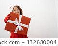 Smiling girl in festive attire presents a red Christmas present, symbolizing holiday generosity and excitement. The white background enhances the joyful and seasonal atmosphere of the image 132680004