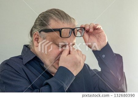 Man in dark shirt adjusts glasses while pinching his nose, showing fatigue and stress 132680390