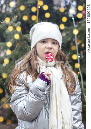 Little girl in winter coat with lollipop near festive tree lights enjoying holiday moment outdoors Little girl in winter coat with lollipop near festive tree lights enjoying holiday moment outdoors 132680408