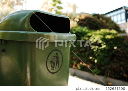 Detailed close-up view of green trash bin with waste icon, located in park area under sunlight. Clean environment and eco-friendly concept 132681608