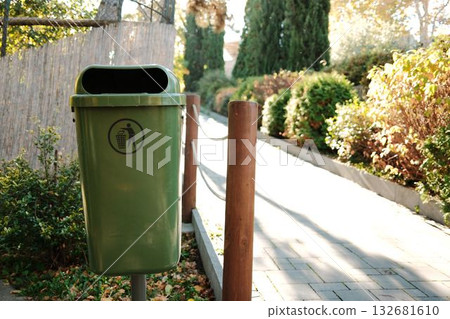 Green trash bin standing beside sunny pathway with plants and trees. Clean environment and eco-friendly concept 132681610