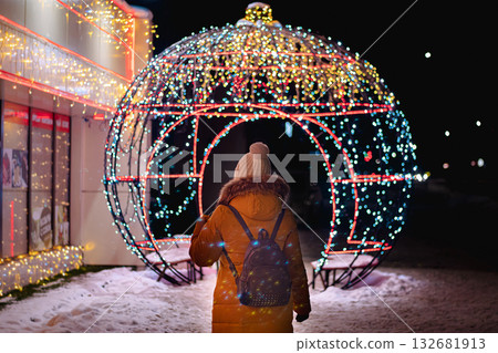 Young woman in knitted hat and warm coat walks through city at night and looks at festive Christmas city decorations. Christmas market, night lights. View from back. Young woman in knitted hat and warm coat walks through city at night and looks at festive Christmas city decorations. Christmas market, night lights. View from back. 132681913