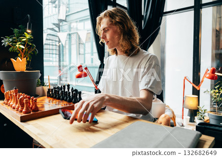 Young man enjoying a chess game in a cozy indoor space with plants Young man enjoying a chess game in a cozy indoor space with plants 132682649