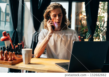 Young man working on his laptop while talking on the phone in a cozy indoor setting Young man working on his laptop while talking on the phone in a cozy indoor setting 132682662