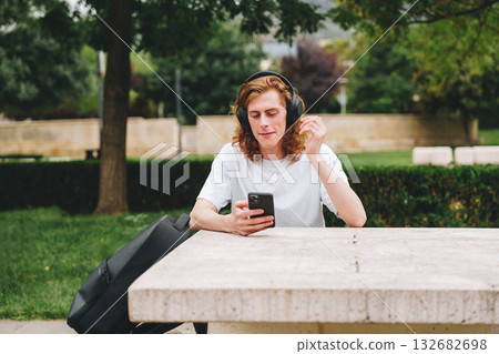 Young man listens to music while using smartphone in park during daytime Young man listens to music while using smartphone in park during daytime 132682698