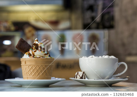 Ice cream dessert in wafer cup with chocolate cookies and and mug of coffee with marshmallows on porcelain plate on blurred colorful copy space background. Ice cream dessert in wafer cup with chocolate cookies and and mug of coffee with marshmallows on porcelain plate on blurred colorful copy space background. 132683468