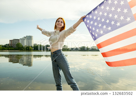Happy young womanl with USA national flag on her shoulders with high city buildings in background celebrating United States independence day. Happy young womanl with USA national flag on her shoulders with high city buildings in background celebrating United States independence day. 132683502