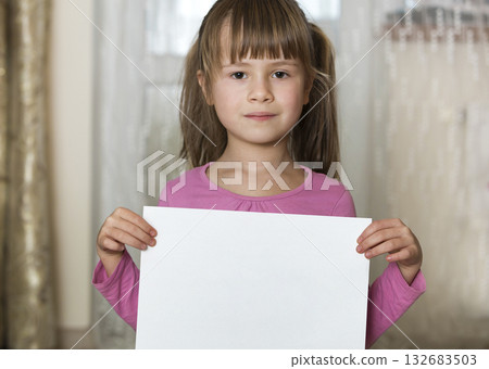 Happy smiling cute child girl holding white copy space sheet of drawing paper. Art education, creativity, advertisement concept. Happy smiling cute child girl holding white copy space sheet of drawing paper. Art education, creativity, advertisement concept. 132683503