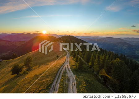 Dirt road winding on hilltop illuminated with bright setting sun. Aerial view of colorful sunset in wild mountains. Beautiful nature landscape 132683670