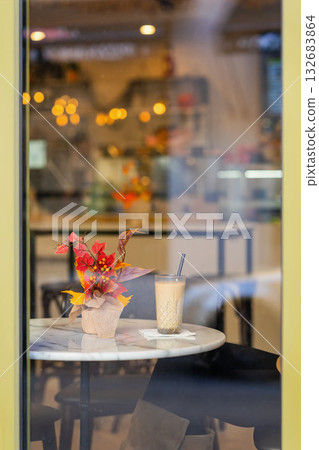 Autumn Decor and Drink in Cafe. Cozy cafe table with autumn flower arrangement and glass of iced coffee behind window reflection. Concept of autumn mood, cafe lifestyle, seasonal vibes Autumn Decor and Drink in Cafe. Cozy cafe table with autumn flower arrangement and glass of iced coffee behind window reflection. Concept of autumn mood, cafe lifestyle, seasonal vibes 132683864