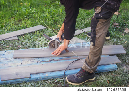 Worker Cutting Tile Outdoors. Construction worker cutting tile with electric grinder on grass field during outdoor renovation project. Concept of craftsmanship, renovation process, skilled labor 132683866
