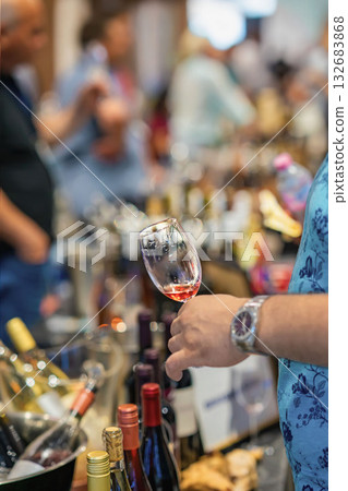 Man holds glass of wine, wine tasting event, surrounded by bottles and people, expo atmosphere. Concept of wine culture, social service gathering 132683868