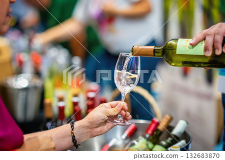 White Wine Being Poured at Tasting Booth. Close-up of white wine being poured into glass, wine tasting event. Hands with glass and bottle, blurred bottles, people in background. Wine expo 132683870