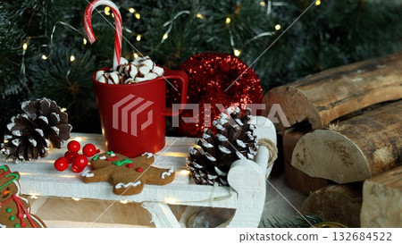 Holiday delight with red mugs of hot chocolate, marshmallows, candy canes, gingerbread cookies, glittering red ornaments, and green pines, embodying joy and cheer of Christmas season. 132684522