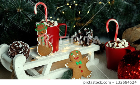 Holiday delight with red mugs of hot chocolate, marshmallows, candy canes, gingerbread cookies, glittering red ornaments, and green pines, embodying joy and cheer of Christmas season. 132684524