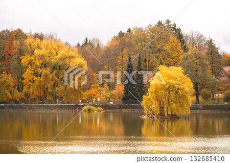 This tranquil pond in Kudowa Zdroj features vibrant autumn colors with shades of orange and yellow. Visitors stroll along the water's edge, enjoying the peaceful fall atmosphere. 132685410