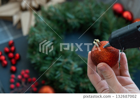 Crafter using hot glue Step by step instruction of making handmade Christmas wreath made of festive decorations in red color. Female hands creating craft homemade decor. New year celebration. Winter Crafter using hot glue Step by step instruction of making handmade Christmas wreath made of festive decorations in red color. Female hands creating craft homemade decor. New year celebration. Winter 132687002