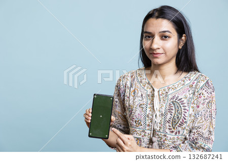 Happy indian woman presenting green screen on her smartphone, posing against the blue background. Joyous girl holding copy space device used for advertising brands in a commercial. 132687241