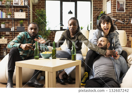 African American man and Caucasian woman compete in video game with wireless controllers while friends watch. Diverse group laughing, bonding and enjoying relaxed hangout in cozy living room. African American man and Caucasian woman compete in video game with wireless controllers while friends watch. Diverse group laughing, bonding and enjoying relaxed hangout in cozy living room. 132687242