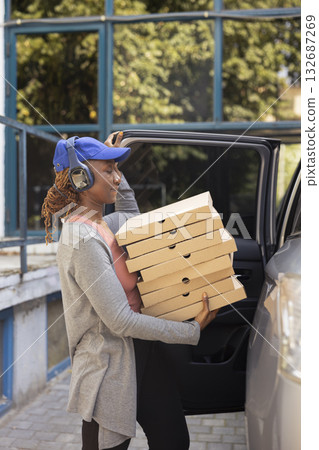 Black takeaway employee parking the car to bring pizza boxes to people at the doorstep, delivering a massive fast food order. Female courier ensuring an express five star service. 132687269
