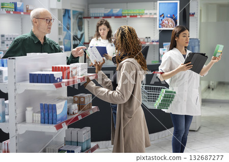 Black young woman helps senior client reading the medication labels, assisting old man in picking up what he needs from the apothecary shelves. Diverse people during pharmacy visit. Black young woman helps senior client reading the medication labels, assisting old man in picking up what he needs from the apothecary shelves. Diverse people during pharmacy visit. 132687277