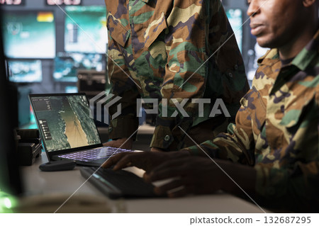 Military officer using laptop device to deploy electronic warfare tools to disrupt enemy communications. Soldier wearing camo uniform operating on high tech equipment helped by notebook 132687295